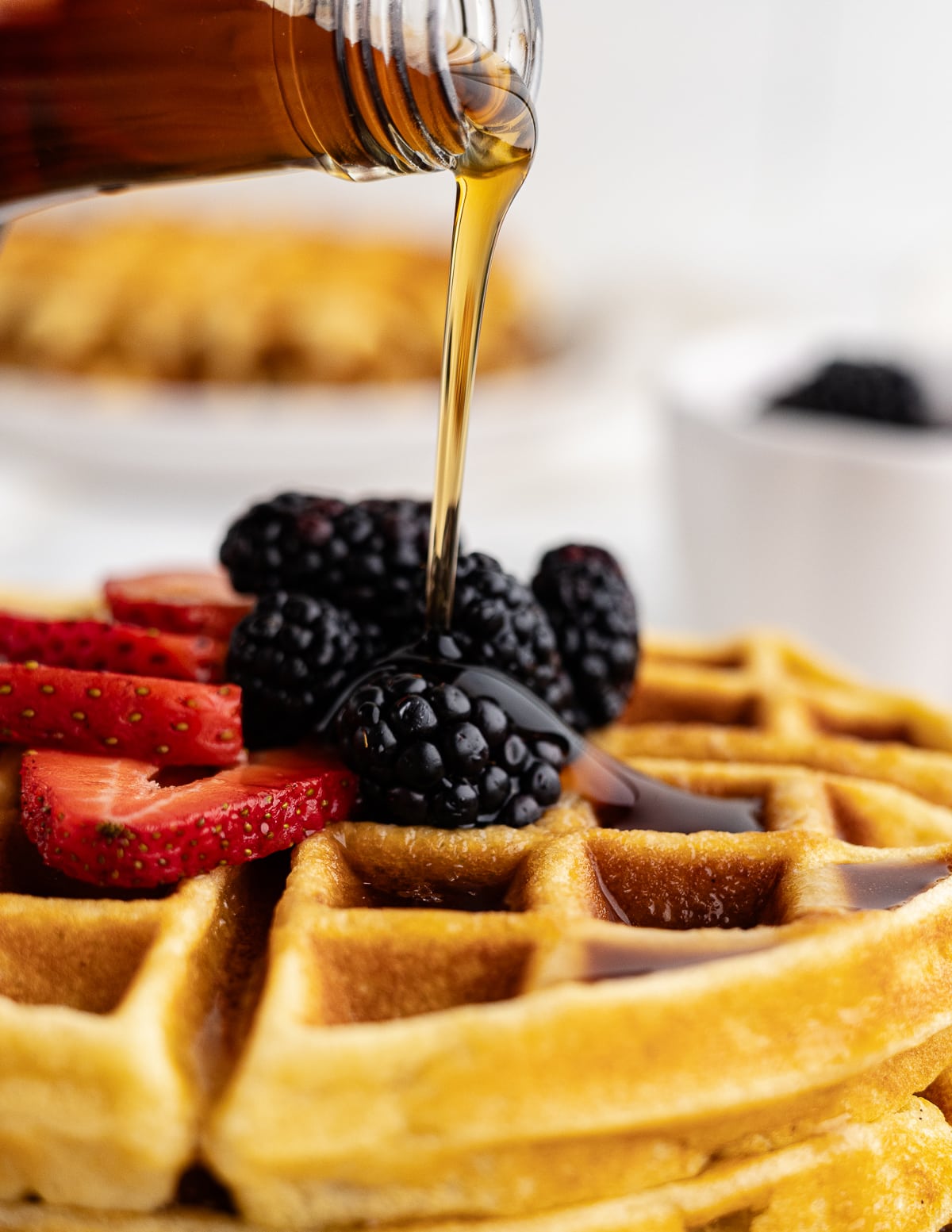 pouring maple syrup onto waffles with blackberries and strawberries on top.