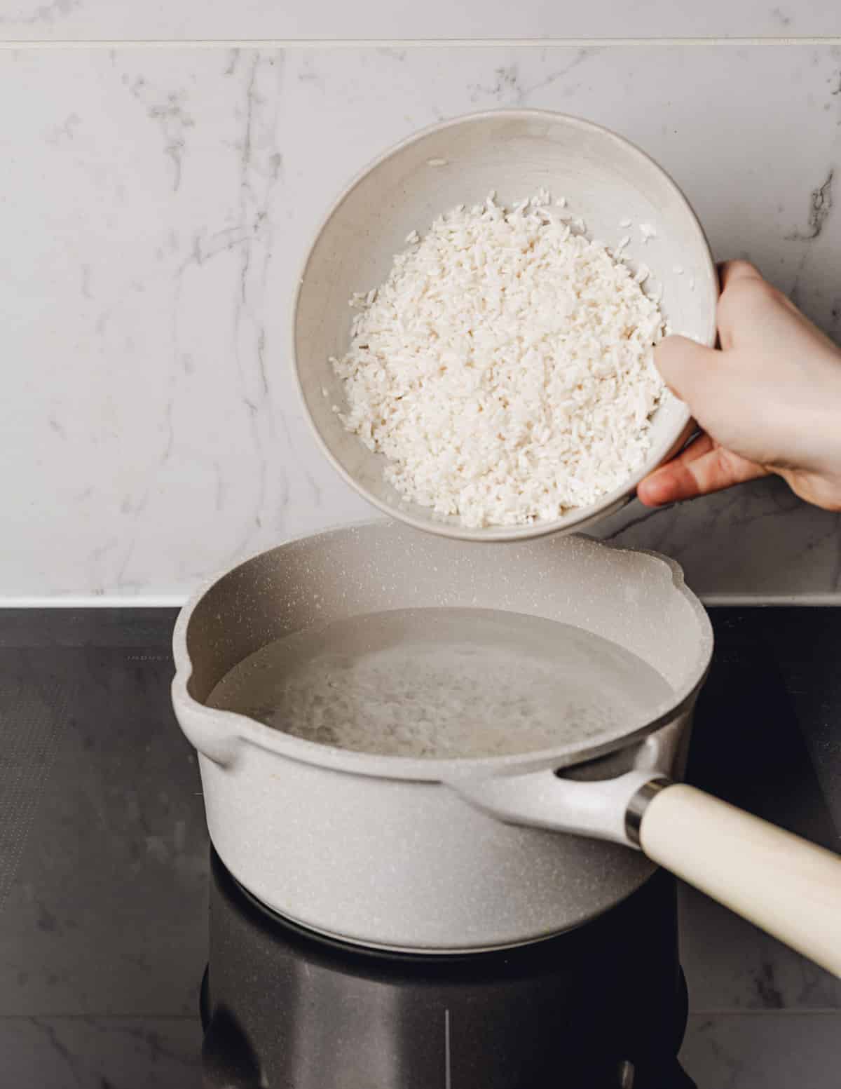 hand pouring a bowl of rice into a pot of boiling water.