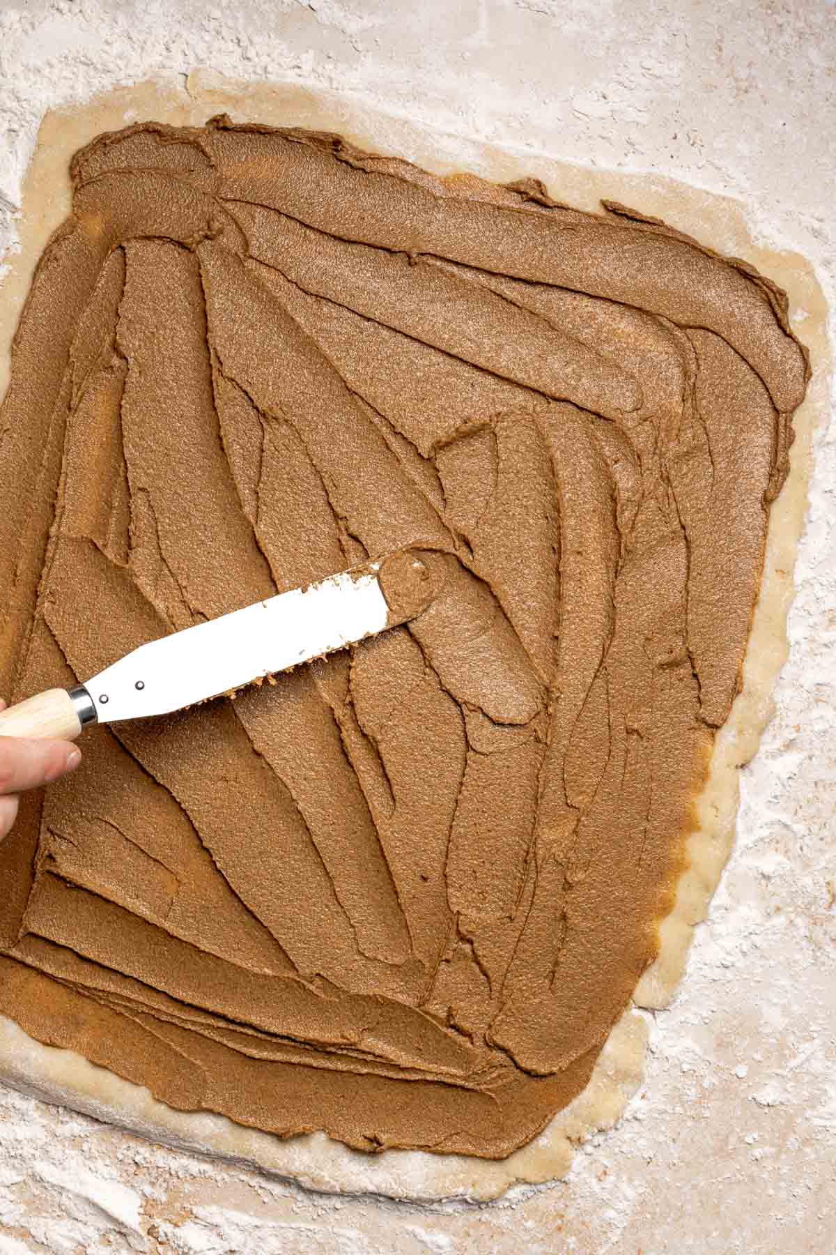 spatula spreading a cinnamon butter mixture onto rolled out dough. 