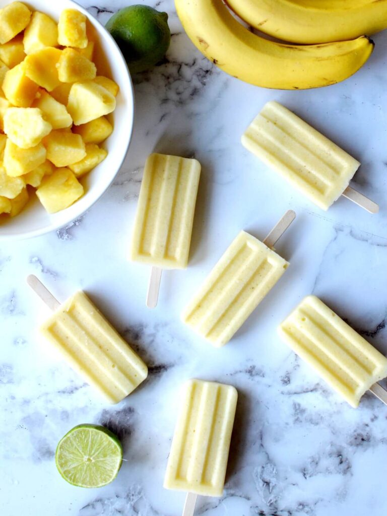 pineapple popsicles laying on a counter with limes, bananas and pineapple around it