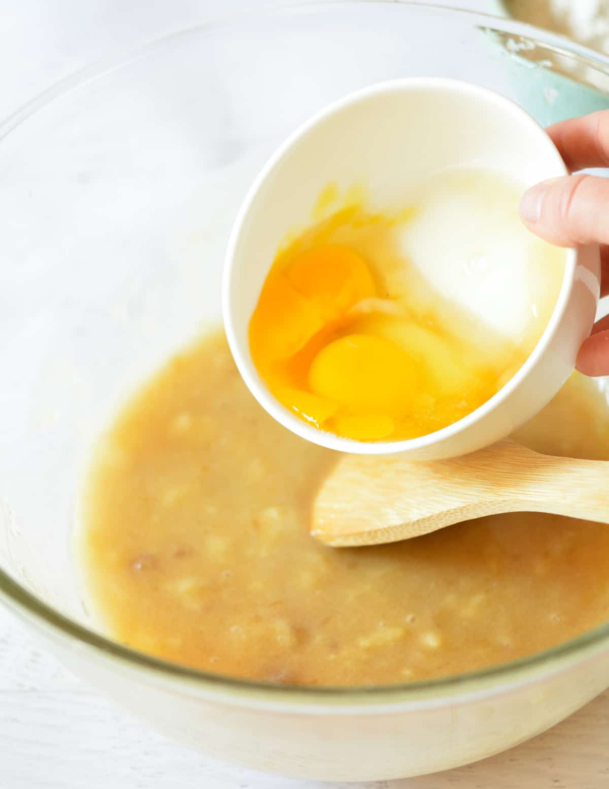 pouring eggs into a bowl of mashed bananas and sugar.