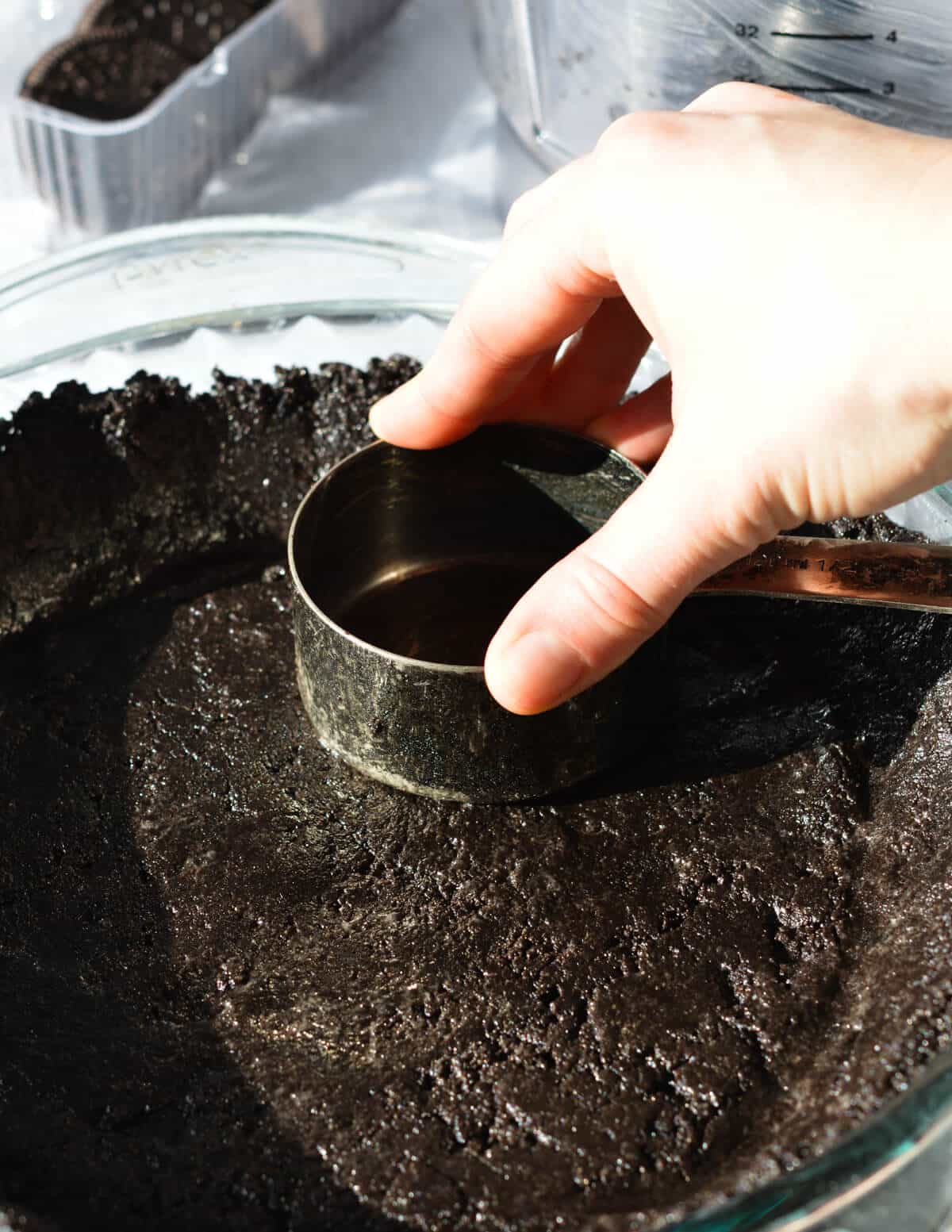 hand pressing a cookie crust into a pan with a measuring cup. 