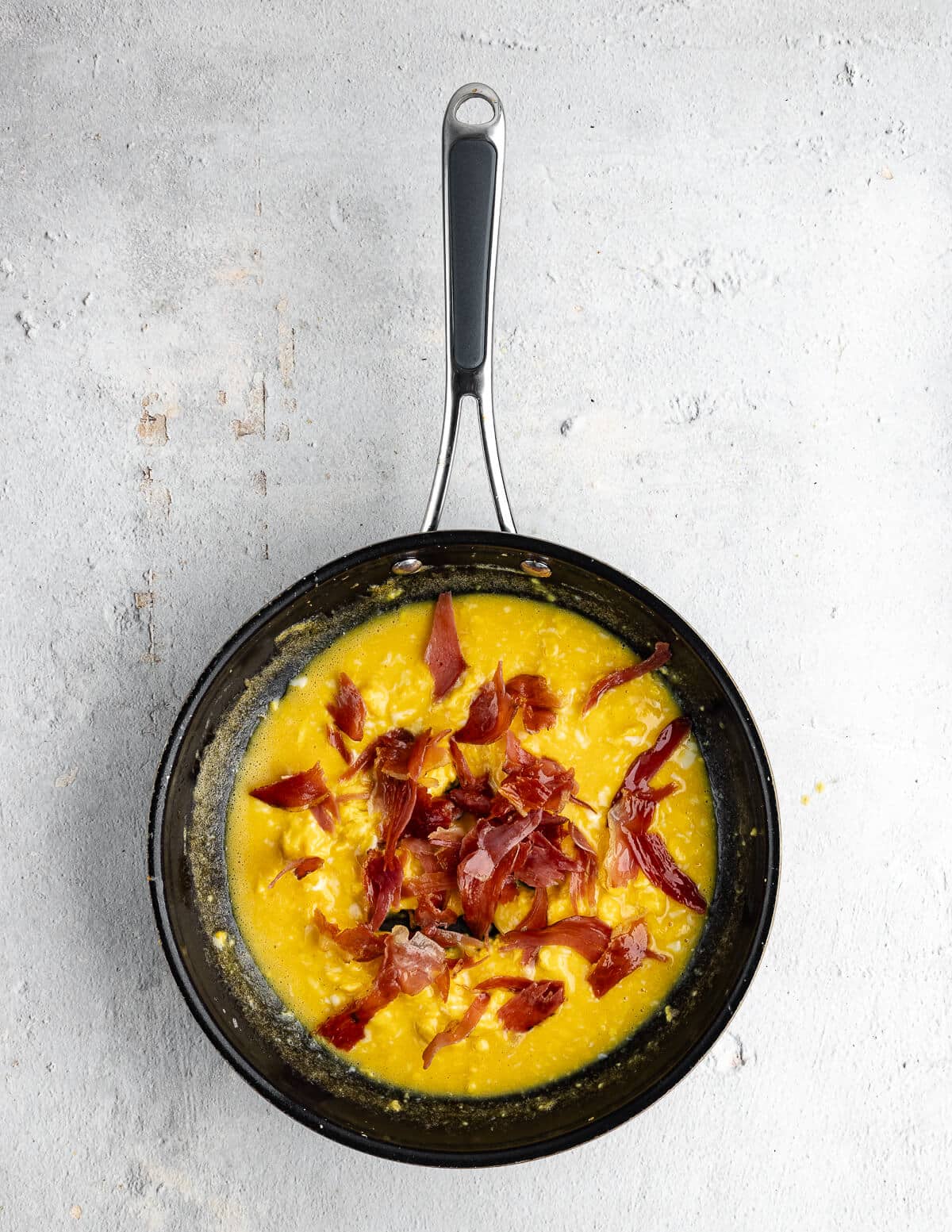 pieces of torn prosciutto over scrambled eggs in a skillet.