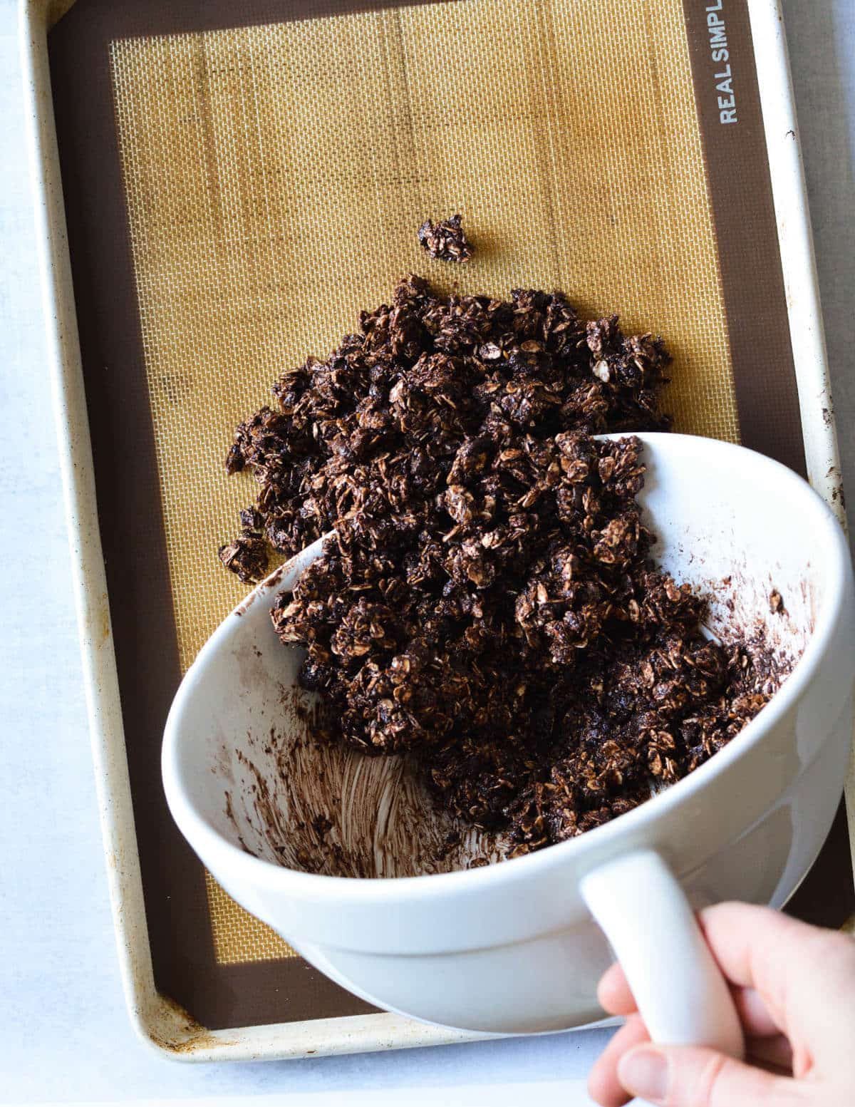 pouring chocolate granola mixture from a bowl to a sheet pan.