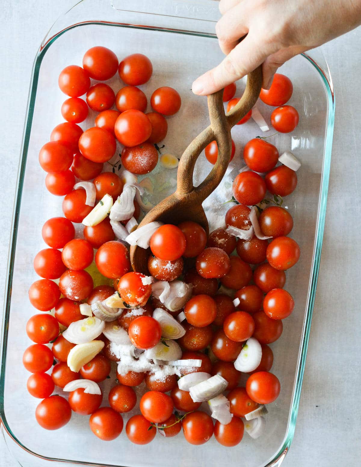 hand with wooden spoon stirring cherry tomatoes, olive oil, shallots, and garlic in a baking dish.