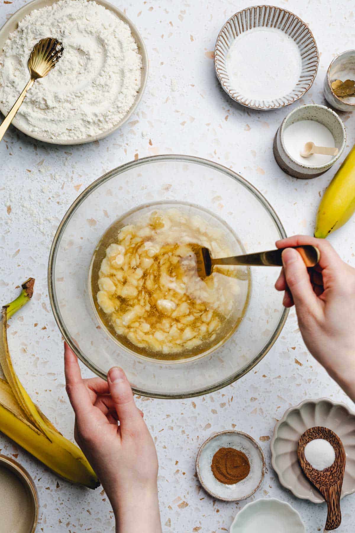 hand using a fork to mix oil, mashed banana, and honey in a bowl.