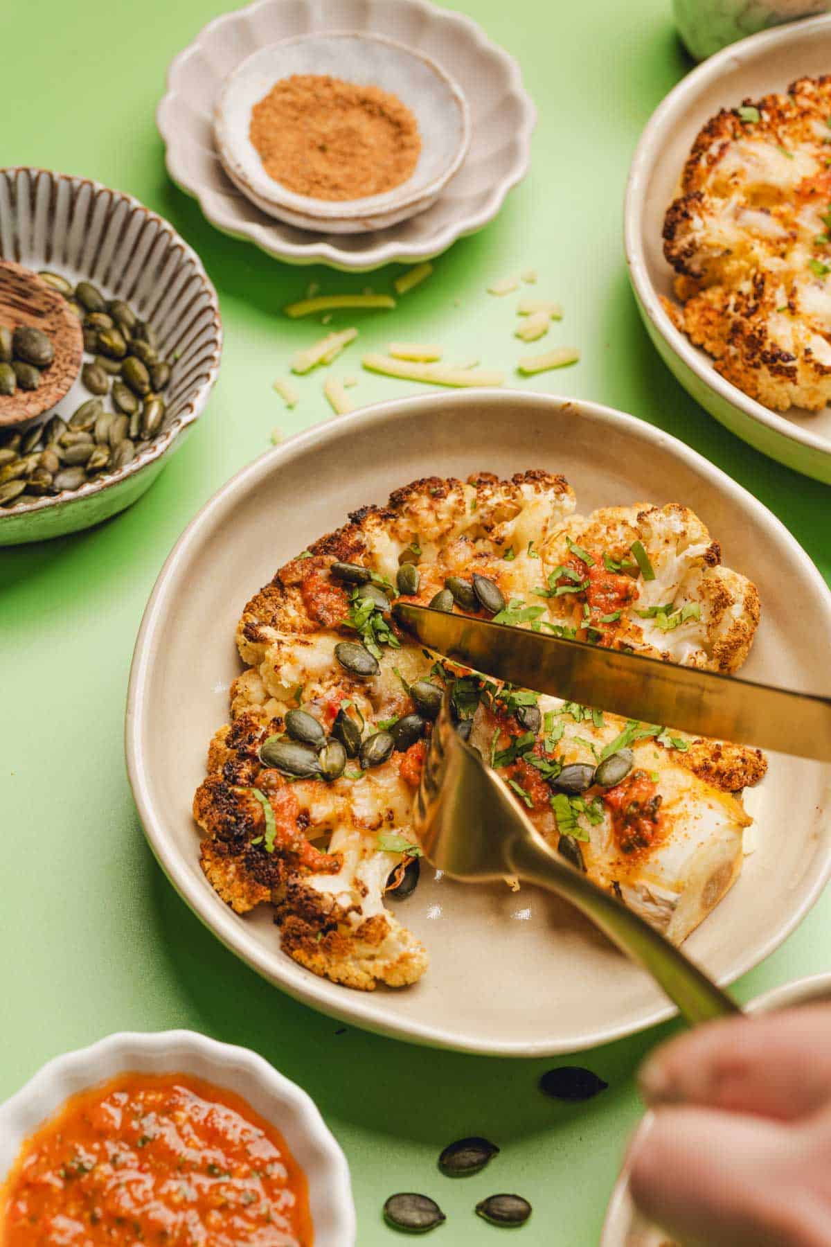 fork and knife cutting into a cheesy cauliflower steak with pepitas and salsa on top.