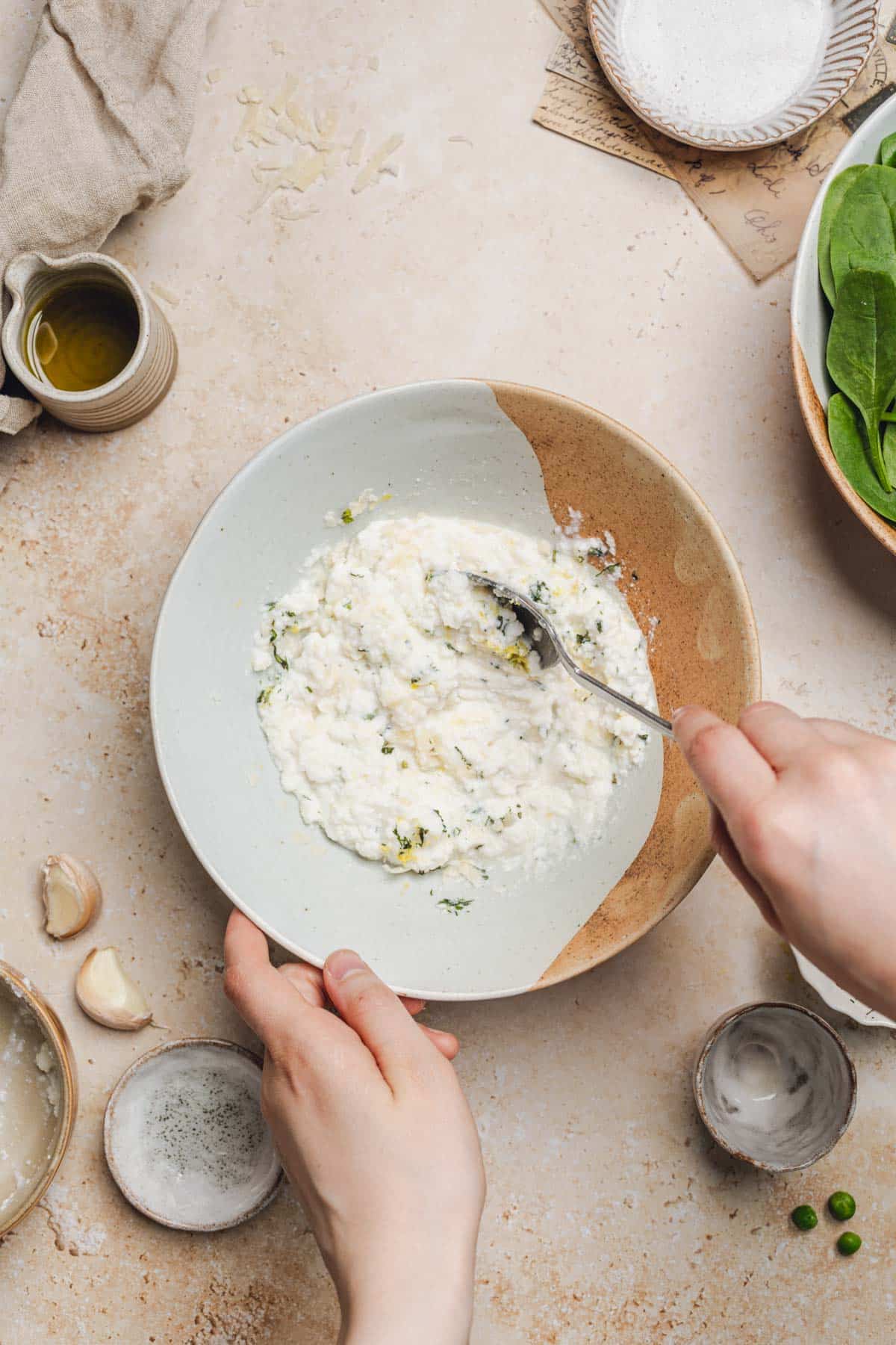 a hand with a spoon mixing ricotta, parmesan, herbs, and lemon juice in a bowl.